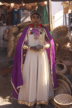 Woman in traditional attire with a purple dupatta and golden dress, standing among woven baskets.